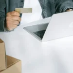 Seseorang menggunakan kartu kredit untuk belanja online di laptop, dikelilingi kotak pengiriman sebagai ilustrasi transaksi e-commerce